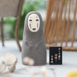 Puede incluir: Una figura de cer&aacute;mica gris oscuro con un cuerpo liso y redondeado y una cara p&aacute;lida de forma ovalada con marcas negras en los ojos y la boca. Al lado hay una peque&ntilde;a placa rectangular negra con texto japon&eacute;s en dorado: 千と千尋の神隠し 信楽焼.