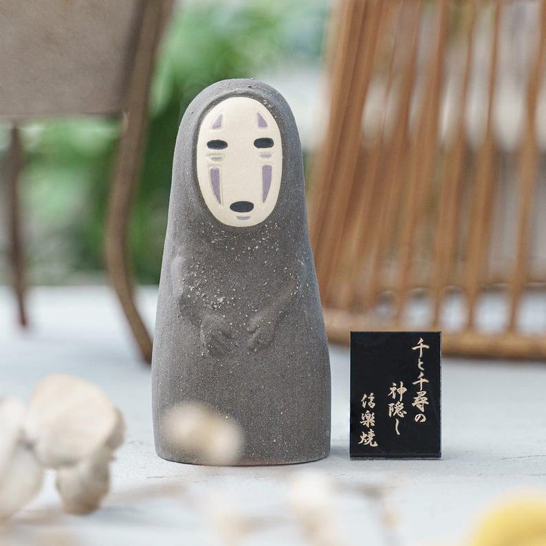Puede incluir: Una figura de cer&aacute;mica gris oscuro con un cuerpo liso y redondeado y una cara p&aacute;lida de forma ovalada con marcas negras en los ojos y la boca. Al lado hay una peque&ntilde;a placa rectangular negra con texto japon&eacute;s en dorado: 千と千尋の神隠し 信楽焼.