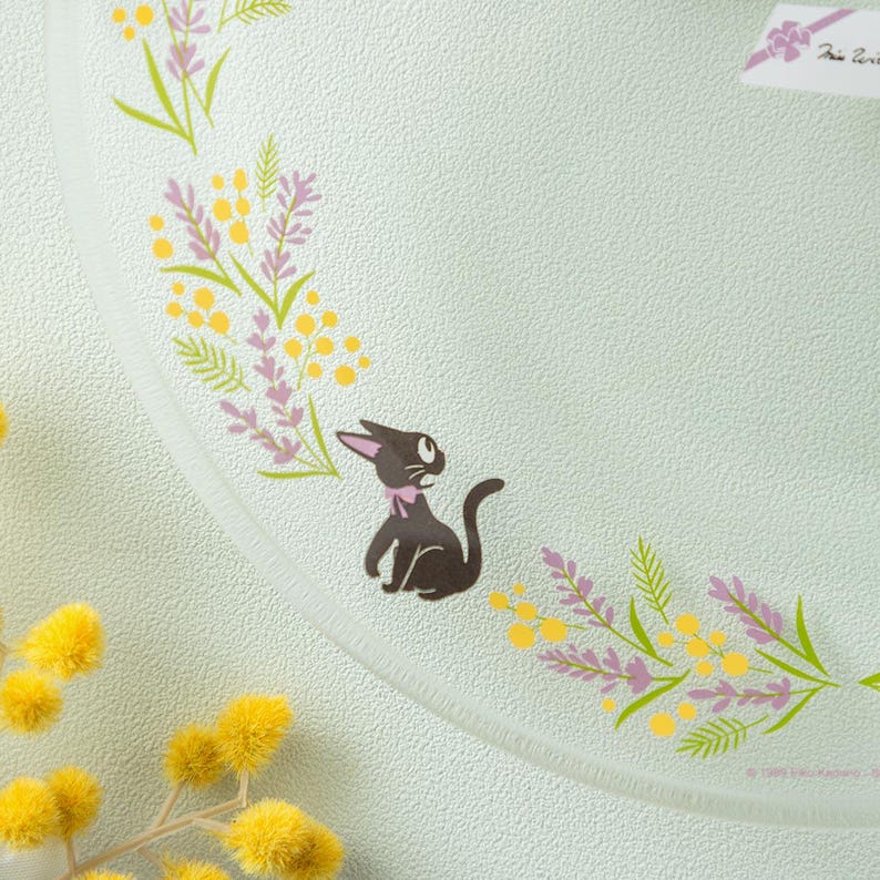May include: A round glass plate with a textured surface featuring a central illustration of a small black cat with a pink bow. The plate is decorated with a curved border of yellow mimosa flowers and purple lavender sprigs. A small white tag is visible.