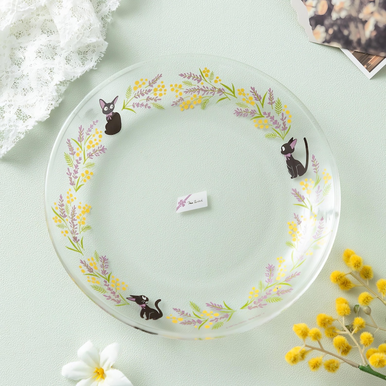 May include: A clear glass plate featuring a decorative border of yellow mimosa flowers and purple lavender sprigs. Three small black cat illustrations are positioned along the floral wreath. A small rectangular white label is centered on the plate.