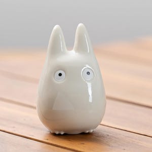K&ouml;nnte beinhalten: Gl&auml;nzende, cremewei&szlig;e Keramikfigur, die an Totoro erinnert. Die Figur hat zwei spitze Ohren, runde Augen und eine dreieckige Nase. Die Figur ist glatt und steht auf einer Holzoberfl&auml;che.