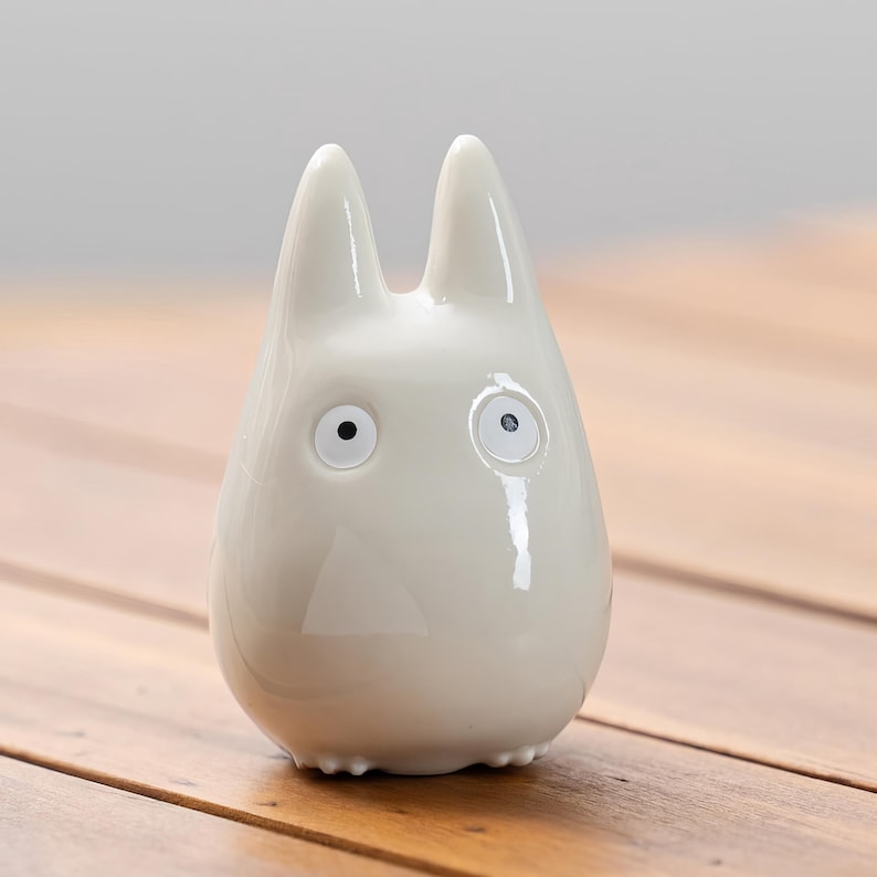 K&ouml;nnte beinhalten: Gl&auml;nzende, cremewei&szlig;e Keramikfigur, die an Totoro erinnert. Die Figur hat zwei spitze Ohren, runde Augen und eine dreieckige Nase. Die Figur ist glatt und steht auf einer Holzoberfl&auml;che.