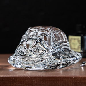 May include: A clear glass turtle figurine with a smiling face and detailed shell. The figurine is sitting on a brown surface, with a dark background. The turtle is a decorative item.