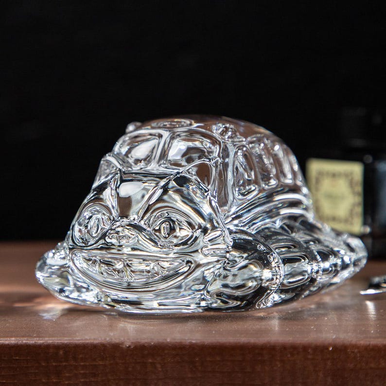 May include: A clear glass turtle figurine with a smiling face and detailed shell. The figurine is sitting on a brown surface, with a dark background. The turtle is a decorative item.