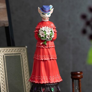Puede incluir: Figura decorativa de un personaje felino con un vestido rojo y un sombrero azul, sosteniendo flores blancas. La figura tiene un dise&ntilde;o detallado con un vestido escalonado y una base decorativa. La base es ornamentada.