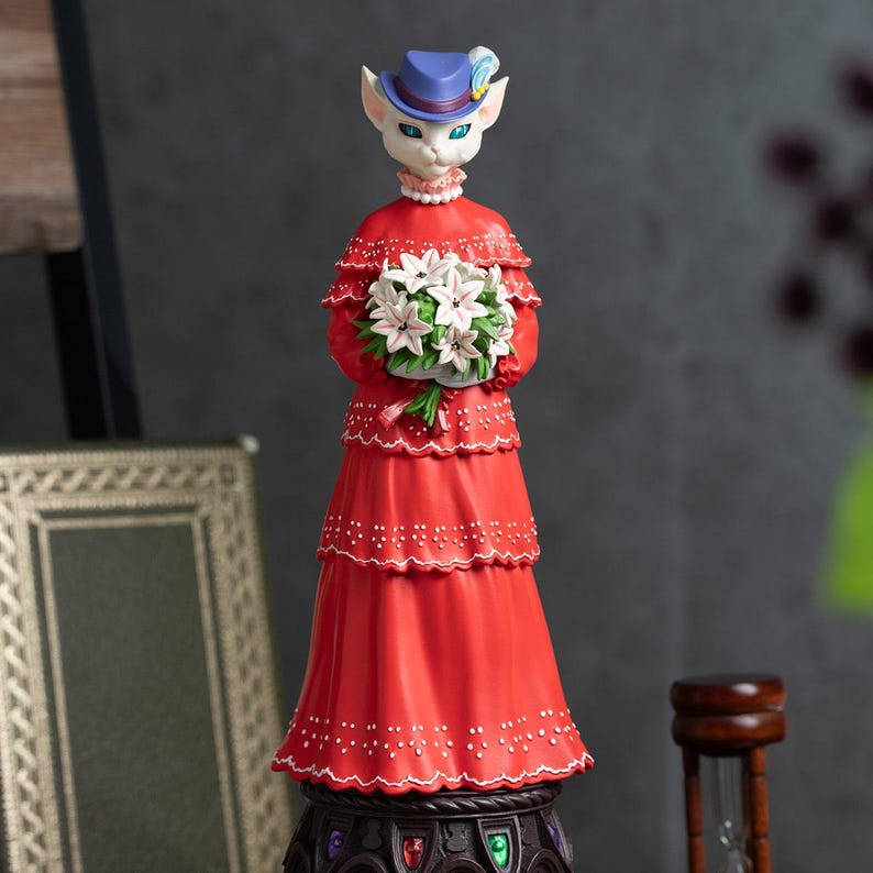 Puede incluir: Figura decorativa de un personaje felino con un vestido rojo y un sombrero azul, sosteniendo flores blancas. La figura tiene un dise&ntilde;o detallado con un vestido escalonado y una base decorativa. La base es ornamentada.