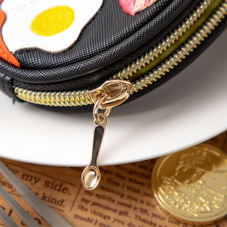 May include: Black pouch with a gold spoon charm and a gold zip. The pouch has embroidered food designs. The background includes a white plate and a gold coin.