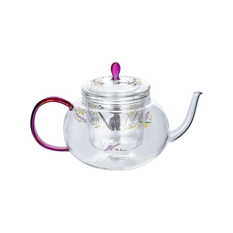 Puede incluir: Una tetera de cristal transparente con un asa morada y un pomo a juego en la tapa. El cuerpo de la tetera est&aacute; decorado con un gr&aacute;fico de un gato negro sentado entre flores amarillas y moradas. Tiene una peque&ntilde;a etiqueta blanca con texto adherida.