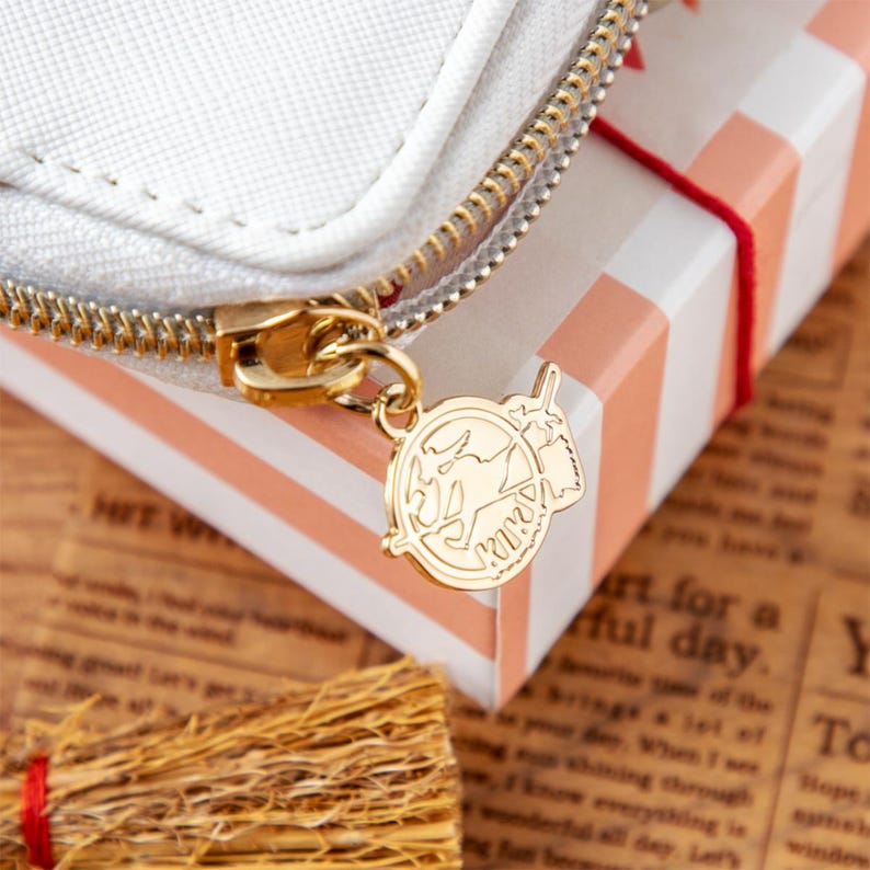May include: Close-up of a white zippered pouch with a gold zipper and charm. The charm features a circular design with a map and a small illustration. The pouch is next to a striped gift box tied with red string.