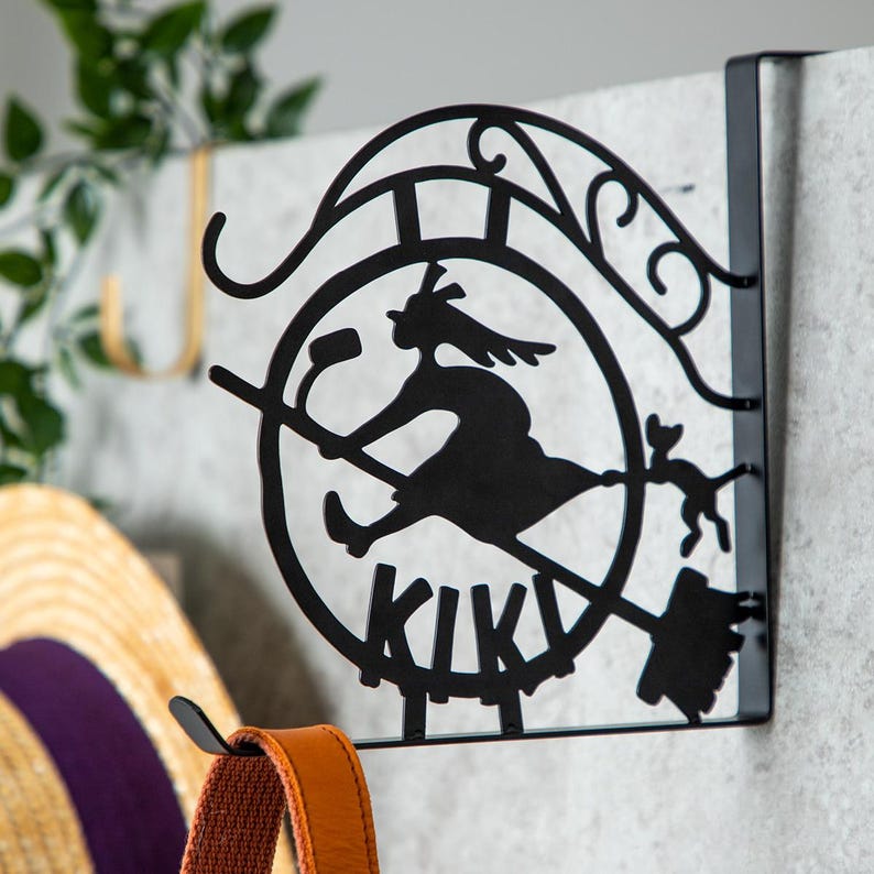 K&ouml;nnte beinhalten: Schwarzer Metall-Wandhaken mit einer Silhouette einer Hexe auf einem Besen mit Katze und dem Wort "KIKI". Der Haken ist zum Aufh&auml;ngen &uuml;ber einer T&uuml;r oder an einer Wand konzipiert.