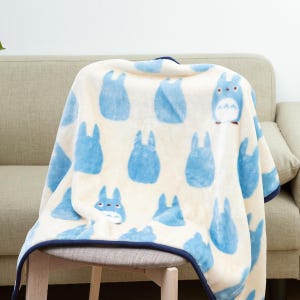 May include: A cream-colored blanket featuring a pattern of blue cartoon characters. The blanket has a dark blue trim and is draped over a chair and sofa. The characters are in various poses.