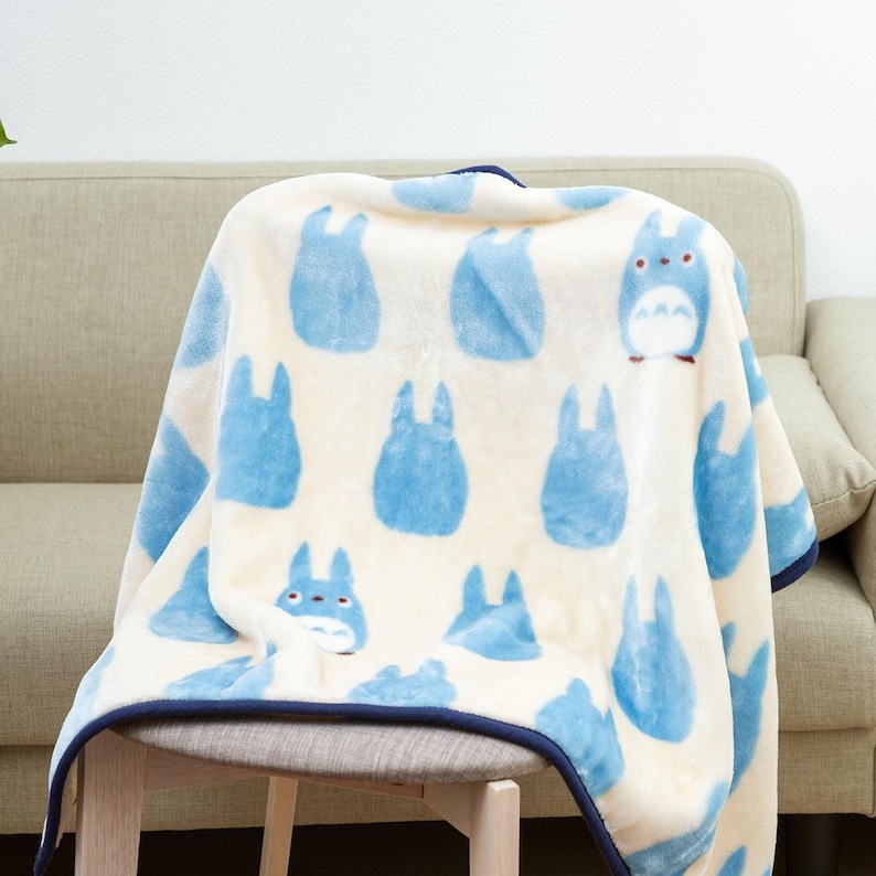 May include: A cream-colored blanket featuring a pattern of blue cartoon characters. The blanket has a dark blue trim and is draped over a chair and sofa. The characters are in various poses.