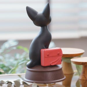 May include: A black cat figurine with a red satchel. The cat is seated on a brown, circular base. The figurine has a sleek, minimalist design, with the cat's ears pointed upwards. The red satchel adds a pop of color to the overall aesthetic.