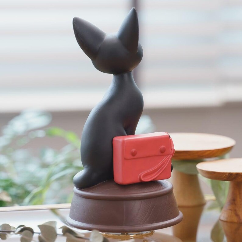 May include: A black cat figurine with a red satchel. The cat is seated on a brown, circular base. The figurine has a sleek, minimalist design, with the cat's ears pointed upwards. The red satchel adds a pop of color to the overall aesthetic.