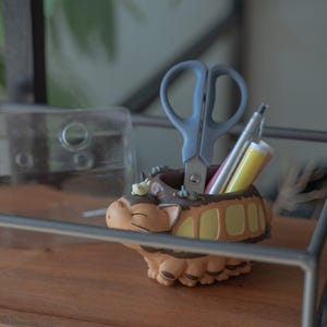 May include: A brown and tan desk organizer shaped like a cartoon character, holding gray scissors, a silver pen, and a yellow and pink cylindrical object. The organizer sits on a wooden surface.