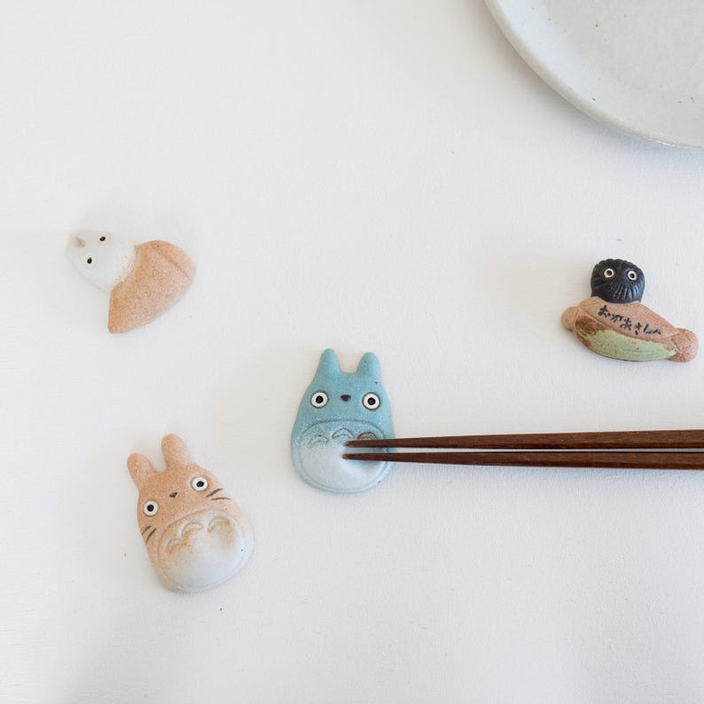 Puede incluir: Cuatro reposa palillos de cer&aacute;mica con figuras estilizadas de animales y criaturas en tonos azul, beige y blanco. Un reposa palillos azul sostiene un par de palillos de madera marr&oacute;n oscuro. Las figuras tienen rasgos faciales pintados.