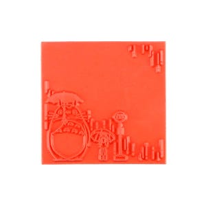 Op de afbeelding: Een vierkante rode rubberen stempel met een verhoogd ontwerp van een groot wezen met een paraplu, een kleiner figuur ernaast met een paraplu en een bushaltebord. Het oppervlak heeft extra verhoogde rechthoekige accenten.