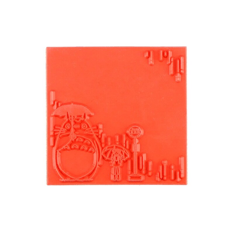 Op de afbeelding: Een vierkante rode rubberen stempel met een verhoogd ontwerp van een groot wezen met een paraplu, een kleiner figuur ernaast met een paraplu en een bushaltebord. Het oppervlak heeft extra verhoogde rechthoekige accenten.