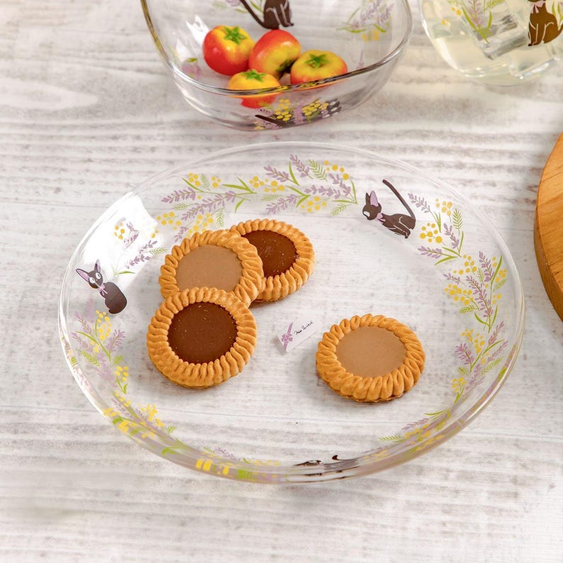 May include: A clear glass plate decorated with a floral pattern of purple and yellow sprigs and black cat silhouettes. Three round cookies with scalloped edges and smooth brown centers are arranged on the plate surface.