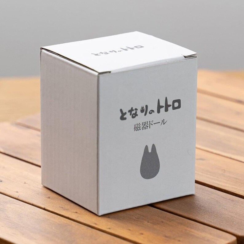 K&ouml;nnte beinhalten: Ein wei&szlig;er Karton mit japanischem Text und einer grauen Silhouette einer Figur. Die Box steht auf einer Holzoberfl&auml;che. Der Text auf der Box lautet "となりのトトロ" und "磁器ドール".
