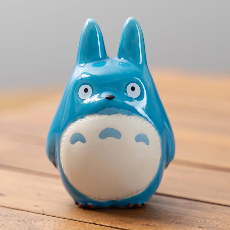 May include: A glossy, turquoise-blue figurine of a cartoon character with large ears, white eyes, and a cream-colored belly with three blue markings. The figurine is set against a blurred background and rests on a wooden surface.