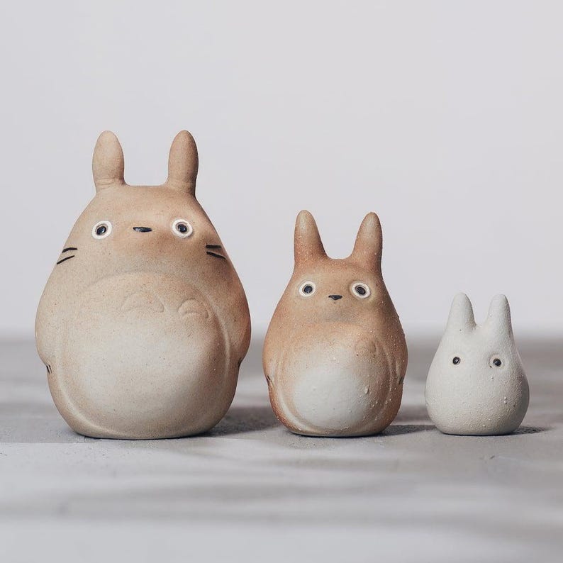 May include: Three ceramic figurines of the character Totoro in varying sizes. The largest is tan with black markings, the middle one is also tan, and the smallest is white. All have large eyes and pointed ears.
