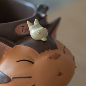 May include: A brown and tan figurine of a character from a Japanese animated film. A small, light-colored creature sits atop the figurine's head. The figurine has a smiling face and the Japanese character "ふ" is visible.