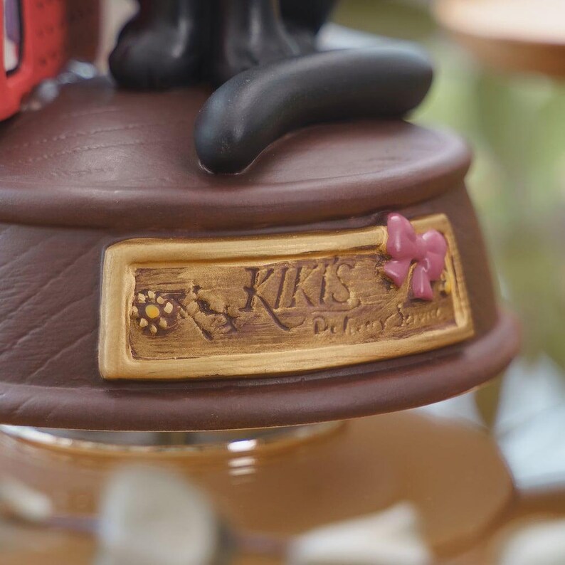 May include: A close-up of a brown decorative base with the text "Kiki's Delivery Service" in gold lettering. A pink bow is on the right side of the base. The base is part of a larger figurine.