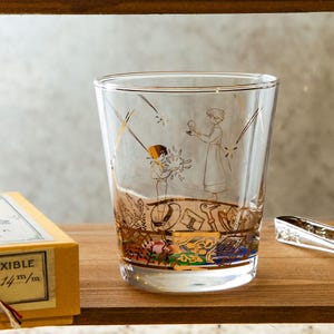 May include: Clear glass tumbler with gold illustrations of figures and objects. The bottom of the glass features a colorful band. The glass is on a wooden surface.
