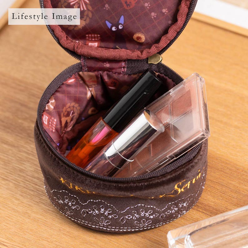 May include: A brown, round cosmetic bag with a zipper, open to reveal makeup items. The bag has a floral patterned interior and a decorative lace design along the bottom. The bag contains lip gloss and an eyeshadow palette.