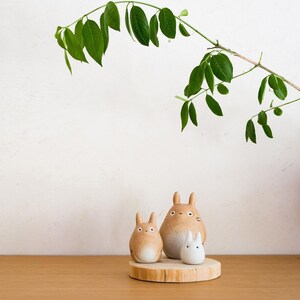 May include: Three ceramic Totoro figurines in varying sizes sit on a wooden slice. The largest is tan with a white belly, the medium one is tan, and the smallest is white. A green leafy branch arches over the figurines.