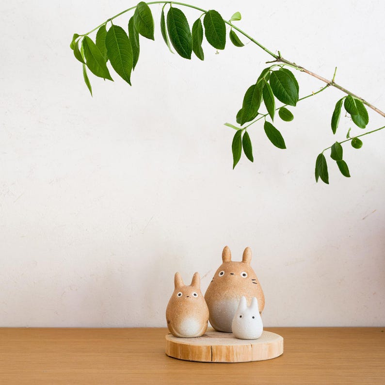 May include: Three ceramic Totoro figurines in varying sizes sit on a wooden slice. The largest is tan with a white belly, the medium one is tan, and the smallest is white. A green leafy branch arches over the figurines.