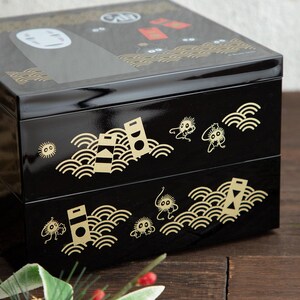 May include: A black, two-tiered bento box with gold accents. The box features illustrations of characters and patterns from a popular animated film. The top tier has a glossy finish, while the lower tiers have a matte finish.
