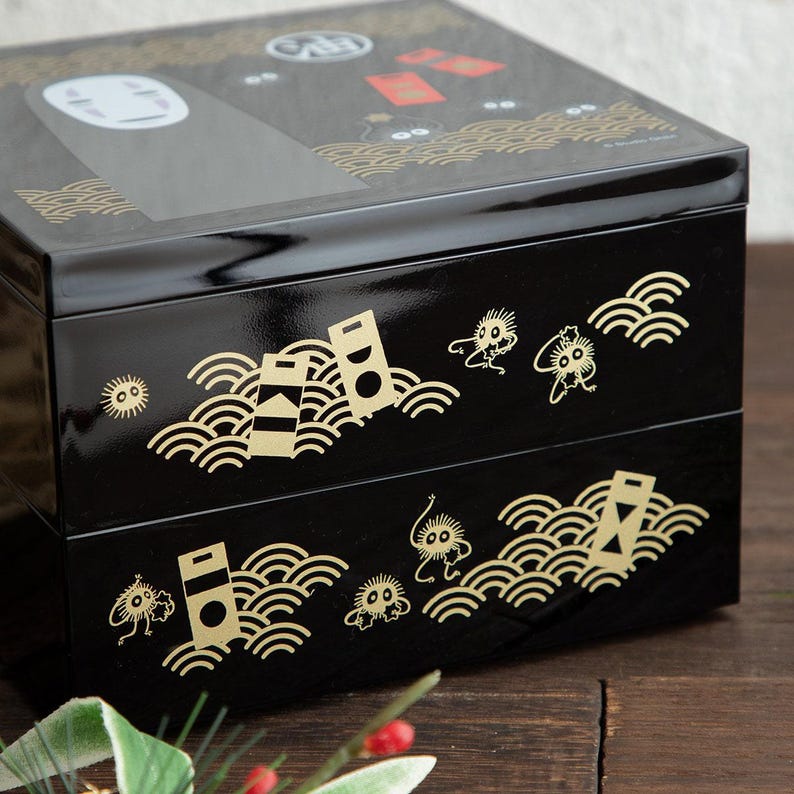 May include: A black, two-tiered bento box with gold accents. The box features illustrations of characters and patterns from a popular animated film. The top tier has a glossy finish, while the lower tiers have a matte finish.