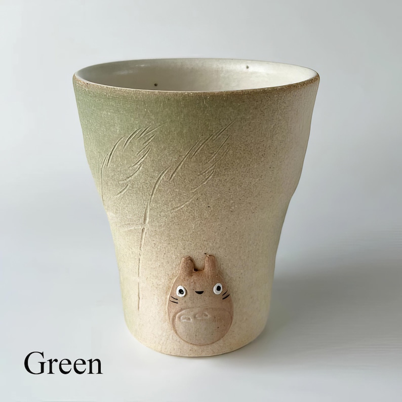 May include: A ceramic tumbler with a light green and beige gradient. The tumbler features an embossed Totoro character and etched leaf designs. The word "Green" is printed at the bottom of the image.