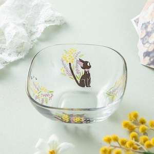 May include: A clear glass bowl featuring a printed graphic of a black cat with a pink bow, surrounded by yellow mimosa flowers and purple lavender sprigs. The bowl has a square shape with rounded corners and a smooth, transparent finish.