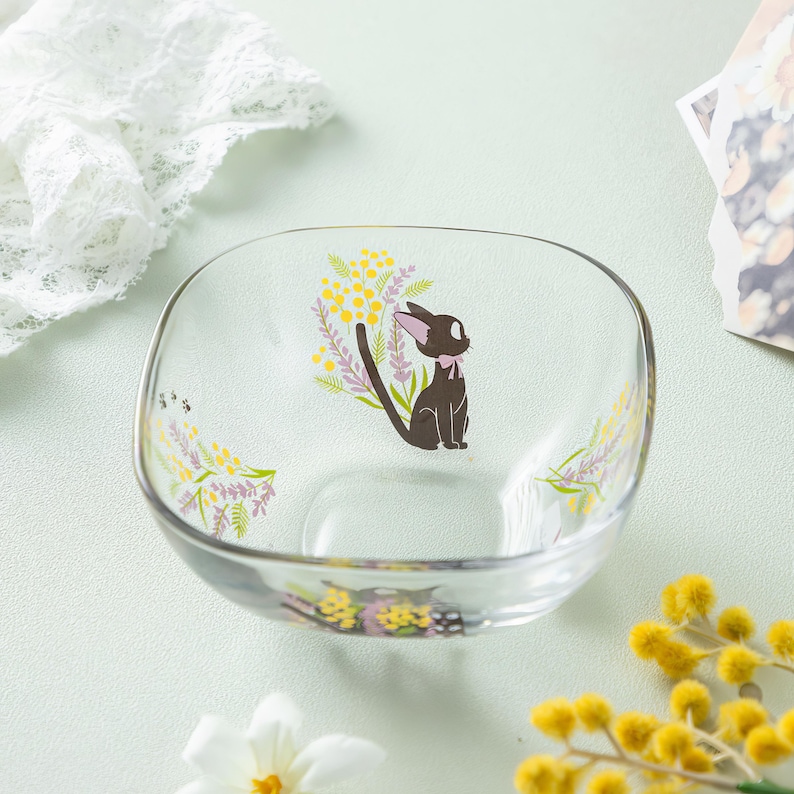 May include: A clear glass bowl featuring a printed graphic of a black cat with a pink bow, surrounded by yellow mimosa flowers and purple lavender sprigs. The bowl has a square shape with rounded corners and a smooth, transparent finish.