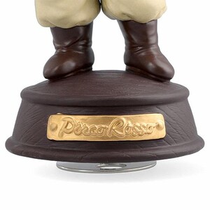 May include: A brown decorative music box base with a gold plaque that reads "Disco Rosso". The base has a textured surface and supports a pair of brown boots and tan trousers.
