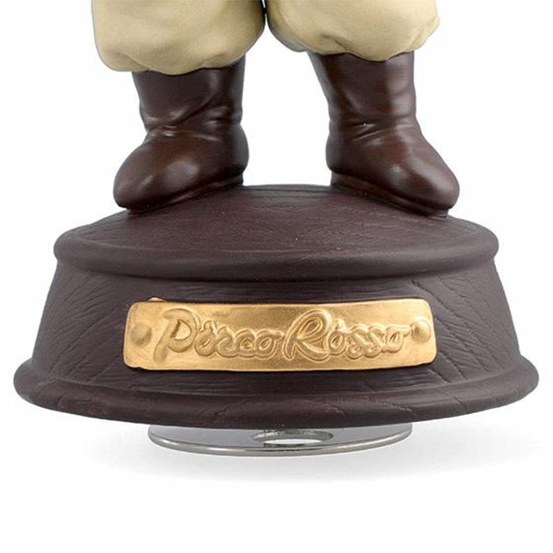May include: A brown decorative music box base with a gold plaque that reads "Disco Rosso". The base has a textured surface and supports a pair of brown boots and tan trousers.