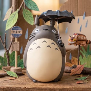 Puede incluir: Una figura de Totoro gris y crema que sostiene un paraguas negro, con un cuerpo redondeado con marcas de chevr&oacute;n en el pecho, orejas puntiagudas y bigotes, situada en una escena con una se&ntilde;al de parada de autob&uacute;s y un peque&ntilde;o veh&iacute;culo felino.