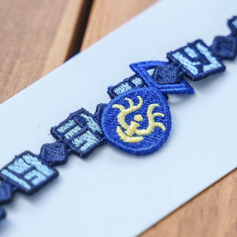 K&ouml;nnte beinhalten: Nahaufnahme eines blau bestickten Accessoires. Das Design zeigt geometrische Formen und ein zentrales Medaillon mit einem gelben Emblem. Das Accessoire liegt auf einer wei&szlig;en Oberfl&auml;che, die die feinen Stickereidetails hervorhebt.