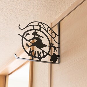 K&ouml;nnte beinhalten: Schwarze Metall-Wanddekoration mit der Silhouette einer Hexe auf einem Besen in einem Kreis, darunter das Wort "KIKI". Das Design umfasst dekorative Elemente und ist an einer Wand befestigt.
