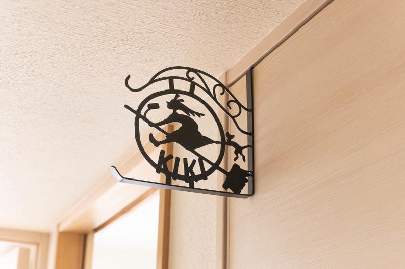 K&ouml;nnte beinhalten: Schwarze Metall-Wanddekoration mit der Silhouette einer Hexe auf einem Besen in einem Kreis, darunter das Wort "KIKI". Das Design umfasst dekorative Elemente und ist an einer Wand befestigt.