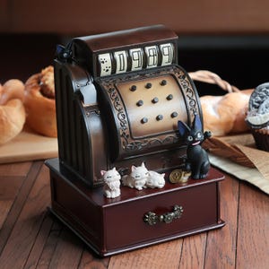 Może przedstawiać: Dekoracyjna kasa fiskalna w stylu vintage, wykończona w kolorze ciemnobrązowym. Wyświetlacz pokazuje "0989". Kasa jest ozdobiona figurkami kot&oacute;w, w tym czarnym kotem i kilkoma białymi kotami. Szuflada jest widoczna na dole.