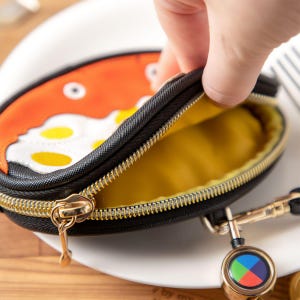 May include: An oval coin purse with a black exterior and a gold zip. The purse has a cartoon fried egg design on the front. The interior is yellow. A retractable keychain is attached. The purse is likely made of fabric.
