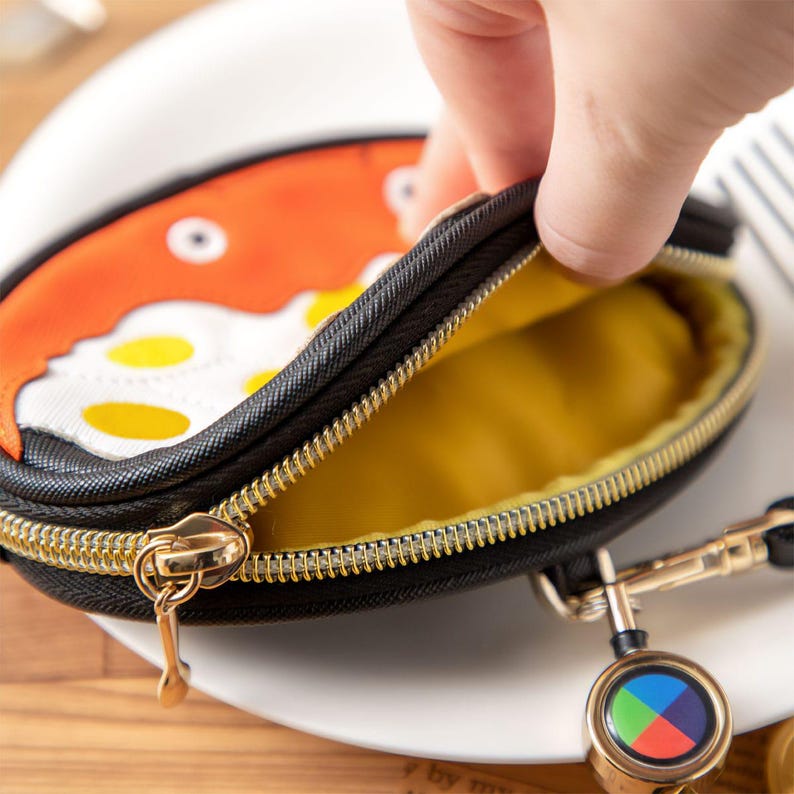 May include: An oval coin purse with a black exterior and a gold zip. The purse has a cartoon fried egg design on the front. The interior is yellow. A retractable keychain is attached. The purse is likely made of fabric.