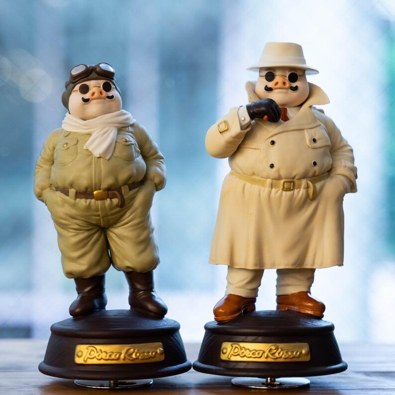 May include: Two pig figurines. One is dressed in a khaki aviator uniform with goggles and a scarf. The other pig is in a cream-coloured trench coat and hat. Both stand on dark brown bases with gold lettering, reminiscent of a film.