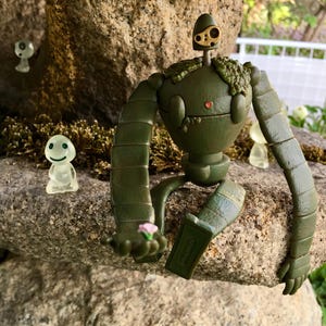 Peut inclure: Figurine de robot vert avec des membres articul&eacute;s, tenant une petite fleur rose. Le robot a une t&ecirc;te ronde avec un petit c&oelig;ur rouge et un dessus dor&eacute;. Deux figurines translucides avec des visages souriants sont &eacute;galement pr&eacute;sentes.