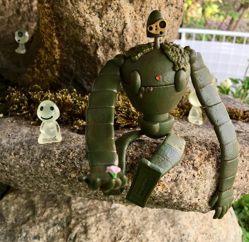 Peut inclure: Figurine de robot vert avec des membres articul&eacute;s, tenant une petite fleur rose. Le robot a une t&ecirc;te ronde avec un petit c&oelig;ur rouge et un dessus dor&eacute;. Deux figurines translucides avec des visages souriants sont &eacute;galement pr&eacute;sentes.