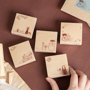May include: Four square wooden rubber stamps featuring detailed brown ink illustrations of animated characters. The designs include a black cat, a large creature with an umbrella at a bus stop, a person with a star, and a hooded figure with a small companion.